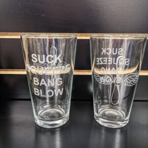 Two Import placeholder for 13627 glasses etched with "SUCK SQUEEZE BANG BLOW," displayed on a black shelf.