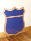 Import placeholder for 410: A blank, shield-shaped blue sign with a tan border, leaning against a wall on wood floor.